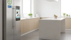 Moderni valoisa keittiö, iso jääkaappi ja valkoinen saareke, kasveja ikkunoilla.